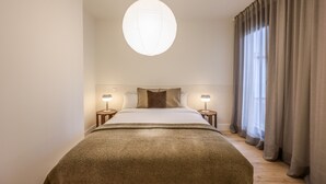Habitació d'hotel moderna amb un llit gran, dues taules de nit amb làmpades, una lluminària penjant i vistes a través de cortines fines.