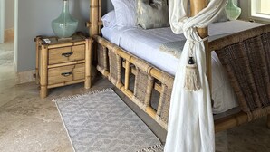 Een bed met hemelbed, een nachtkastje met lamp en een geweven vloerkleed op een betegelde vloer.