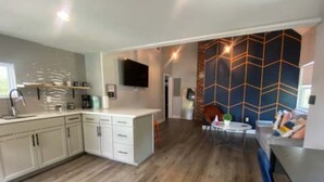 A modern kitchen with white cabinets, a built-in oven, and a coffee machine. There is a dining area with a table and chairs, and a living room with a sofa and a TV mounted on the wall. - The Contemporary (Sanford)