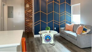 A modern living room with a geometric-patterned wall, a grey sofa with colorful cushions, a round coffee table, and a brick accent wall. - The Contemporary (Sanford)