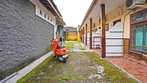 A narrow alleyway with a scooter parked on the left, a building with a tiled floor on the right, and a visible air conditioning unit.