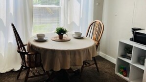 A dining area with a round table, four chairs, a potted plant, and a white shelf with items. - Corporate Rental Apartment In Northwest Indiana (Lake Station)