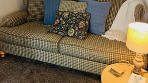 A checkered sofa with floral and solid pillows, a round wall clock, a lamp, and a small table with a tissue box. - Corporate Rental Apartment In Northwest Indiana (Lake Station)