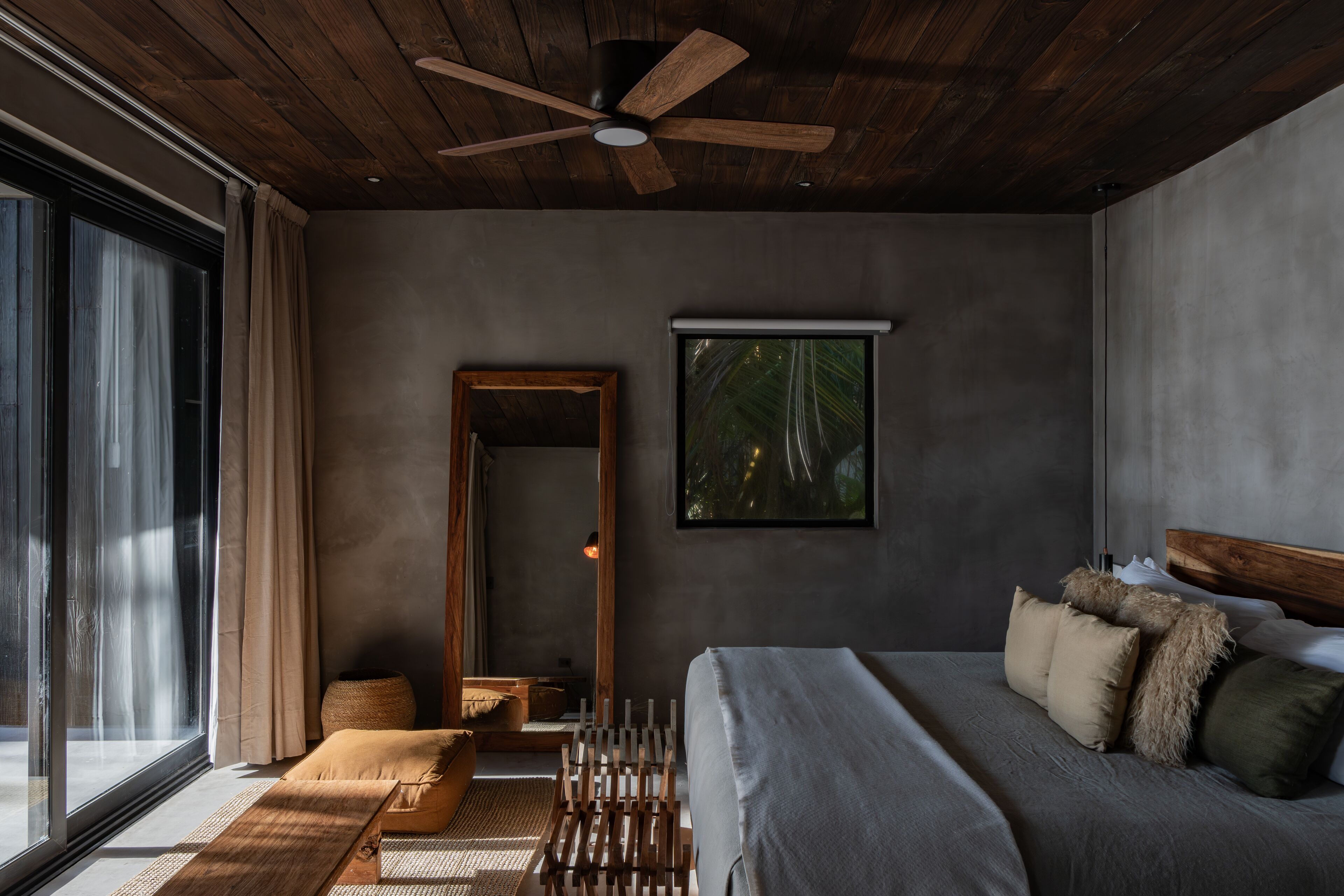 A modern bedroom with a large bed, wooden ceiling fan, and a view of the outdoors through a large window.