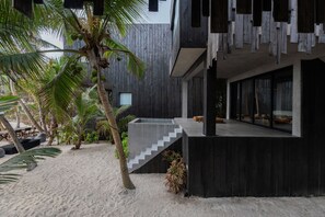 A modern building with a concrete staircase leading to a balcony, surrounded by palm trees and a sandy area.