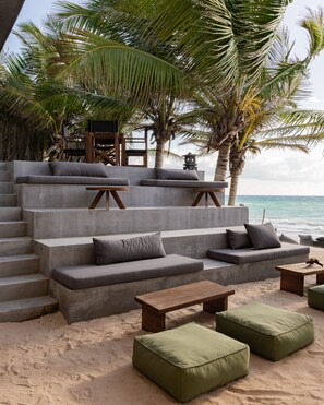 A beachside lounge with concrete steps leading to a seating area, surrounded by palm trees and overlooking the ocean.