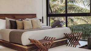 Een bed met een houten hoofdbord, meerdere kussens en uitzicht op palmbomen door grote ramen.