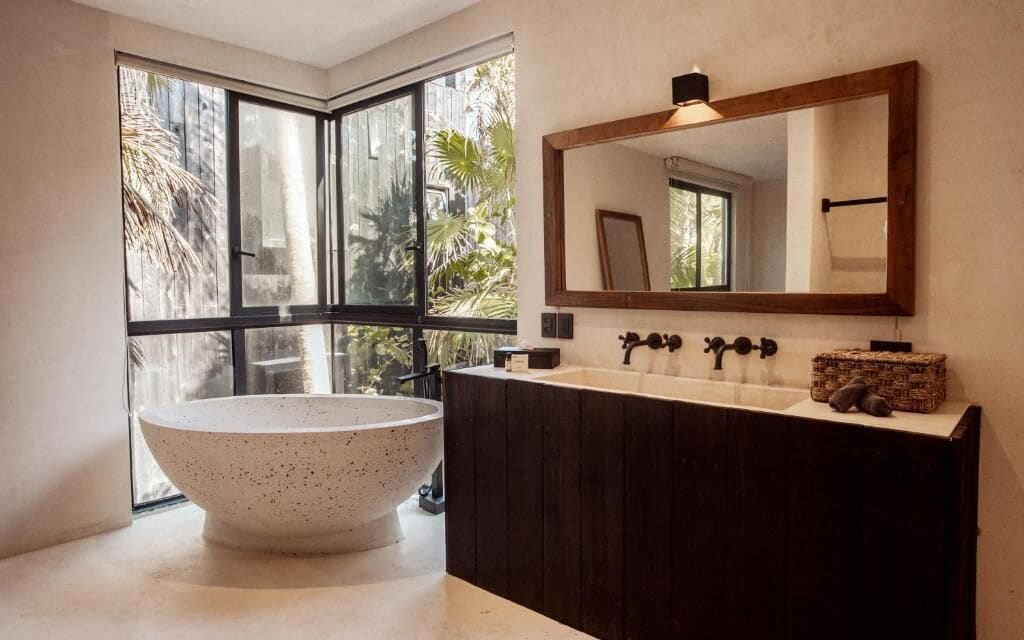 Un baño moderno con una bañera independiente, un espejo grande y una ventana con vistas a palmeras.