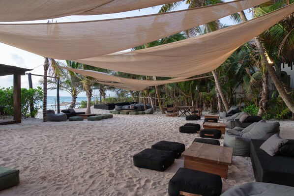 A beachside lounge with outdoor seating, including sofas and bean bags, under a canopy of shade.