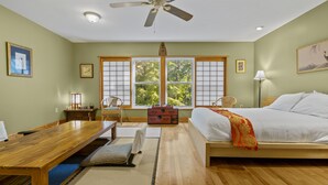 Une chambre avec un lit en bois, un bureau, une chaise, une fenêtre donnant sur des arbres et un ventilateur de plafond.