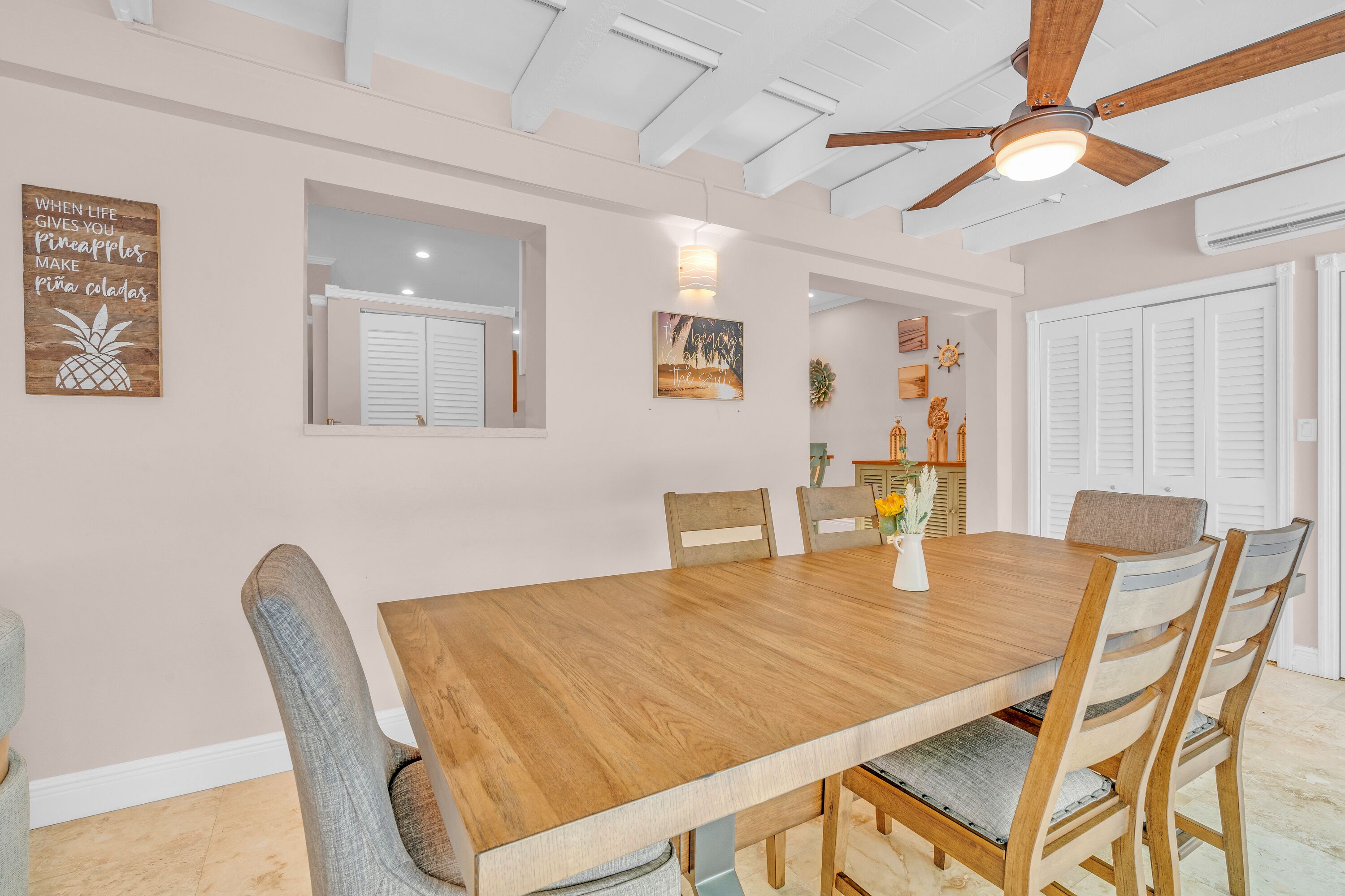 A dining area with a wooden table and chairs, a ceiling fan, and wall decorations including a pineapple sign and a framed picture.