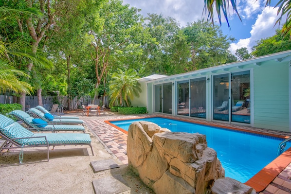 A backyard with a swimming pool, lounge chairs, and a small building with large windows.