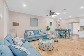 A living room with a sofa, ottoman, dining table, chairs, ceiling fan, and a plant.