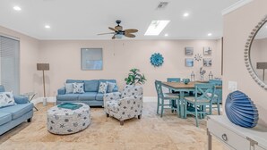 Un salon avec un canapé bleu, un pouf à motifs et un coin repas comprenant une table et des chaises en bois.