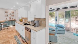 Une cuisine moderne avec des meubles blancs, un lave-vaisselle et une machine à café. On y trouve également un coin repas avec une table et des chaises, ainsi qu’une vue sur une piscine et des chaises longues à l’extérieur.