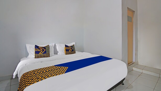 A neatly made bed with a blue and yellow patterned bedspread and pillows, a white wall, and a door leading to another room. - SPOT ON 92498 Fanni Homestay (Pekanbaru)