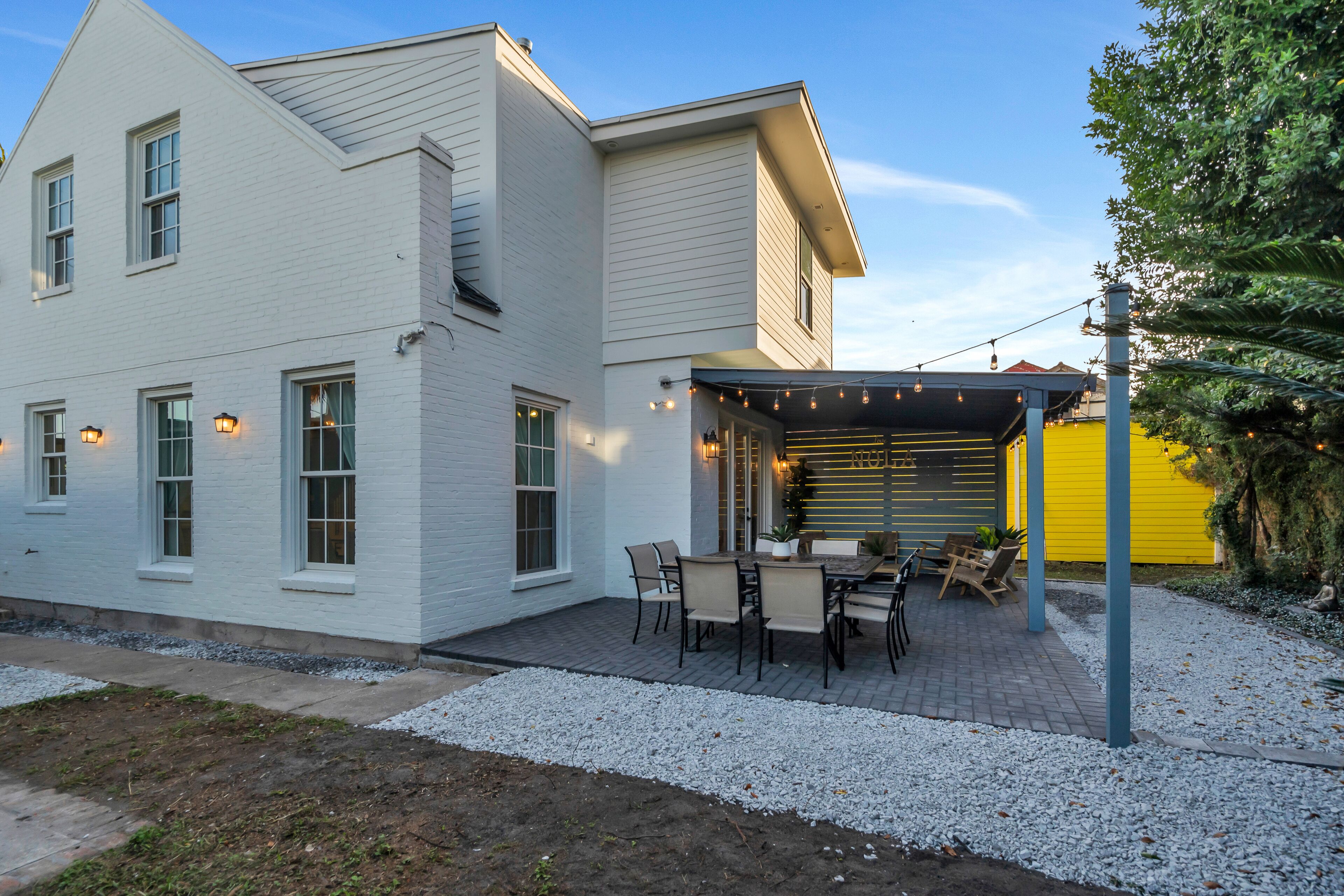 A white two-story house with a patio area featuring a table and chairs, string lights, and a yellow wall.