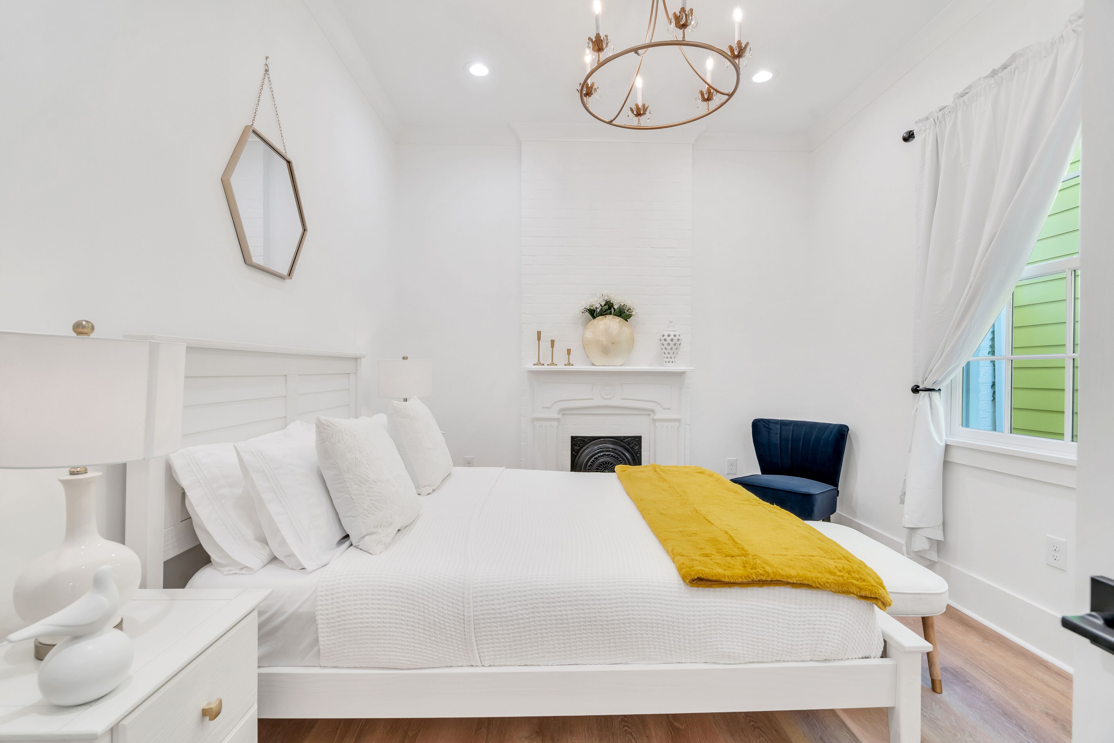 A white bedroom with a bed, bedside tables, a mirror, a chandelier, a fireplace, and a window with curtains.