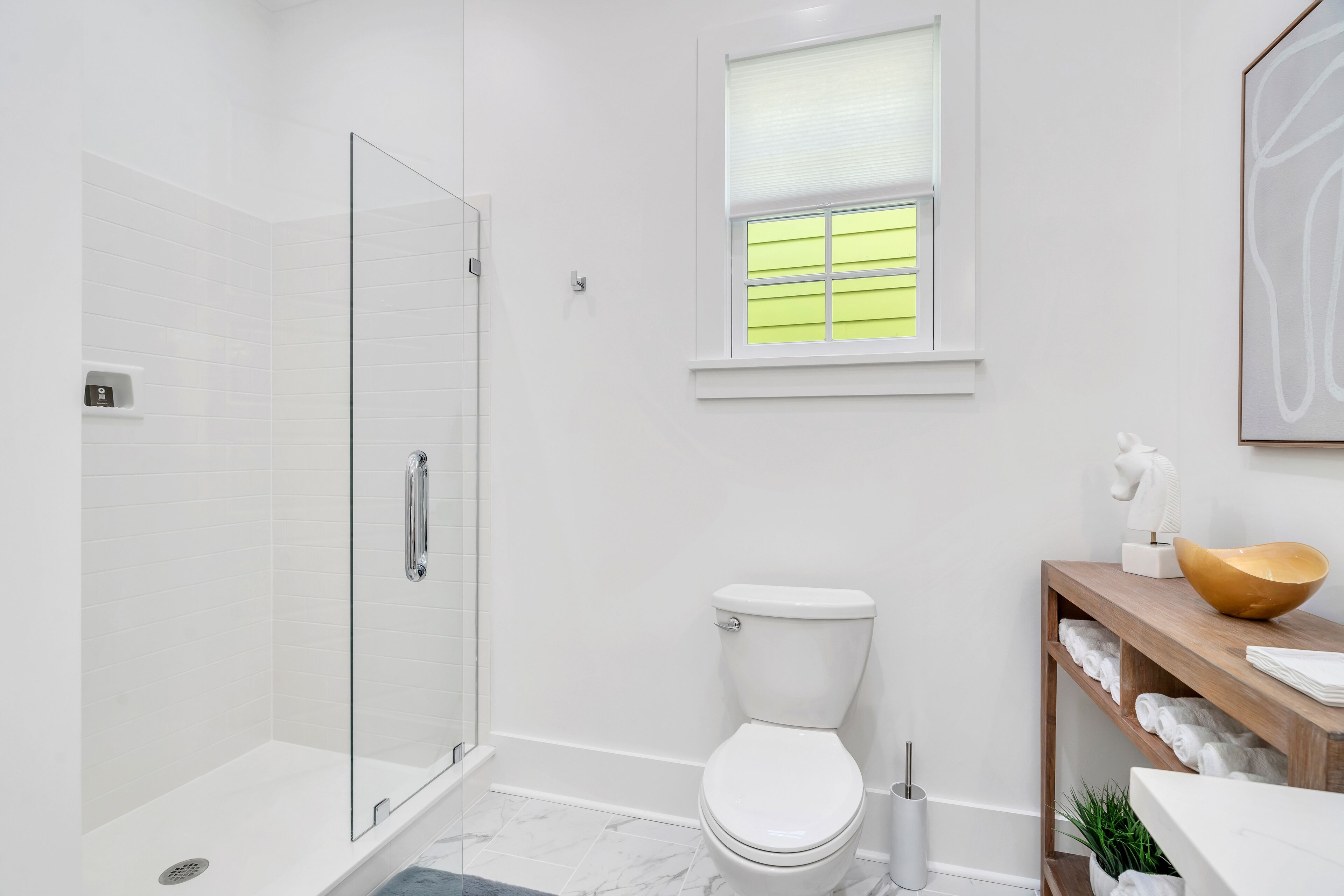 Un baño moderno con cabina de ducha de vidrio, inodoro, ventana con persianas y tocador de madera con toallas.