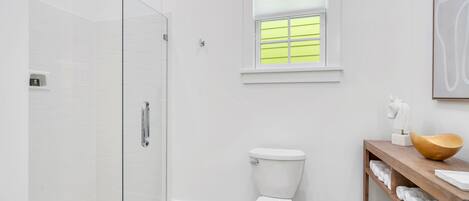 Un baño moderno con cabina de ducha de vidrio, inodoro, ventana con persianas y tocador de madera con toallas.