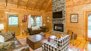 A cozy log cabin interior with a stone fireplace, wooden ceiling beams, and a comfortable seating area.