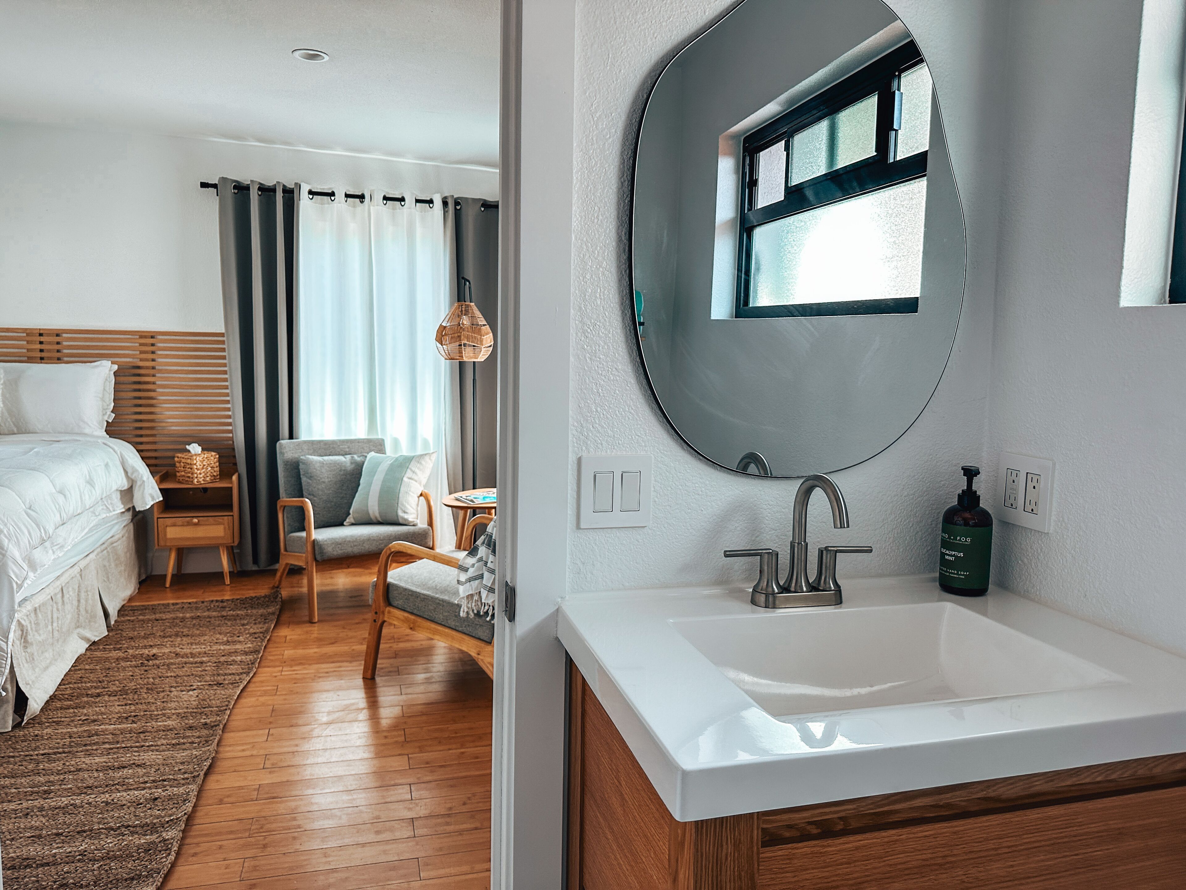 A hotel room with a bed, a chair, a small table, a round mirror, and a white sink.