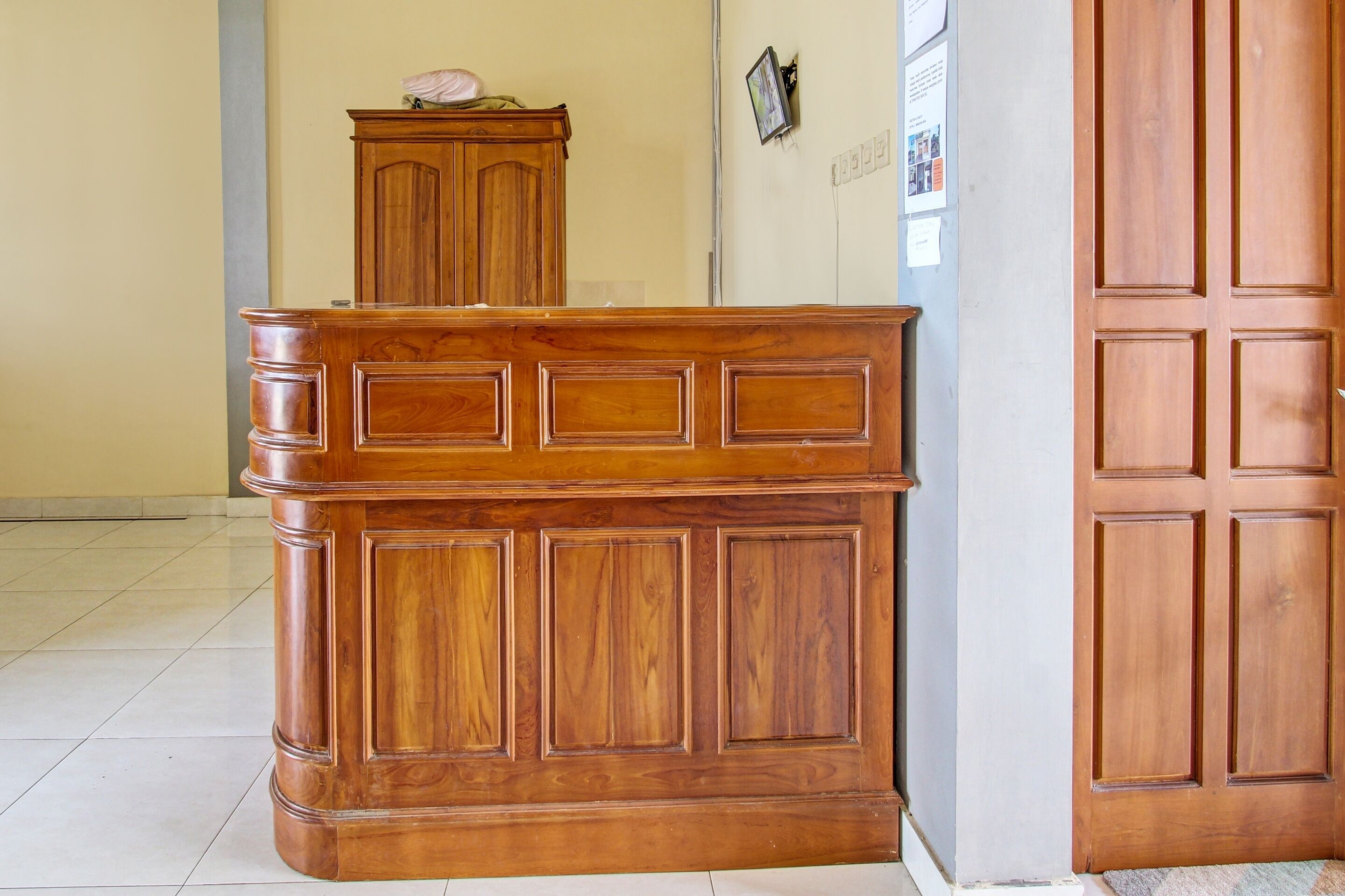 A wooden reception desk with a cabinet above it, a door to the right, and a wall with a light switch and a small picture.