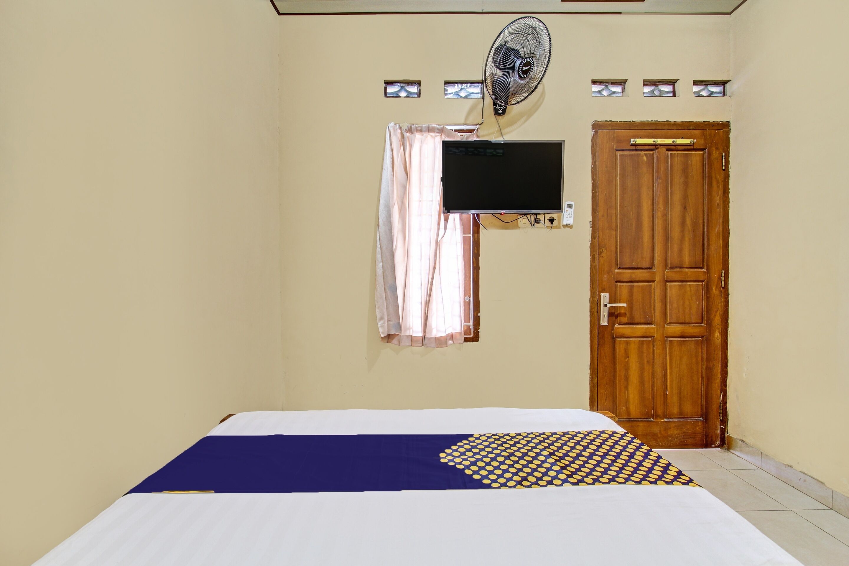 Una habitación con una cama, un televisor montado en la pared, una puerta de madera y un ventilador.