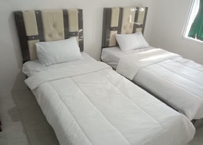 Duas camas de solteiro com roupa de cama branca num quarto com chão em azulejos e janela com cortina verde.