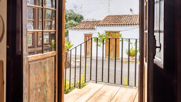 Une porte en bois ouverte donne sur un balcon d’où l’on peut admirer un bâtiment blanc avec un toit recouvert de tuiles et une clôture en métal.