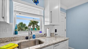 A kitchen with a granite countertop, white cabinets, a stainless steel sink, a window with blinds, and a view of palm trees outside. - Surfs Up - 1 Block to Beach & Walk to Dining (Flagler Beach)