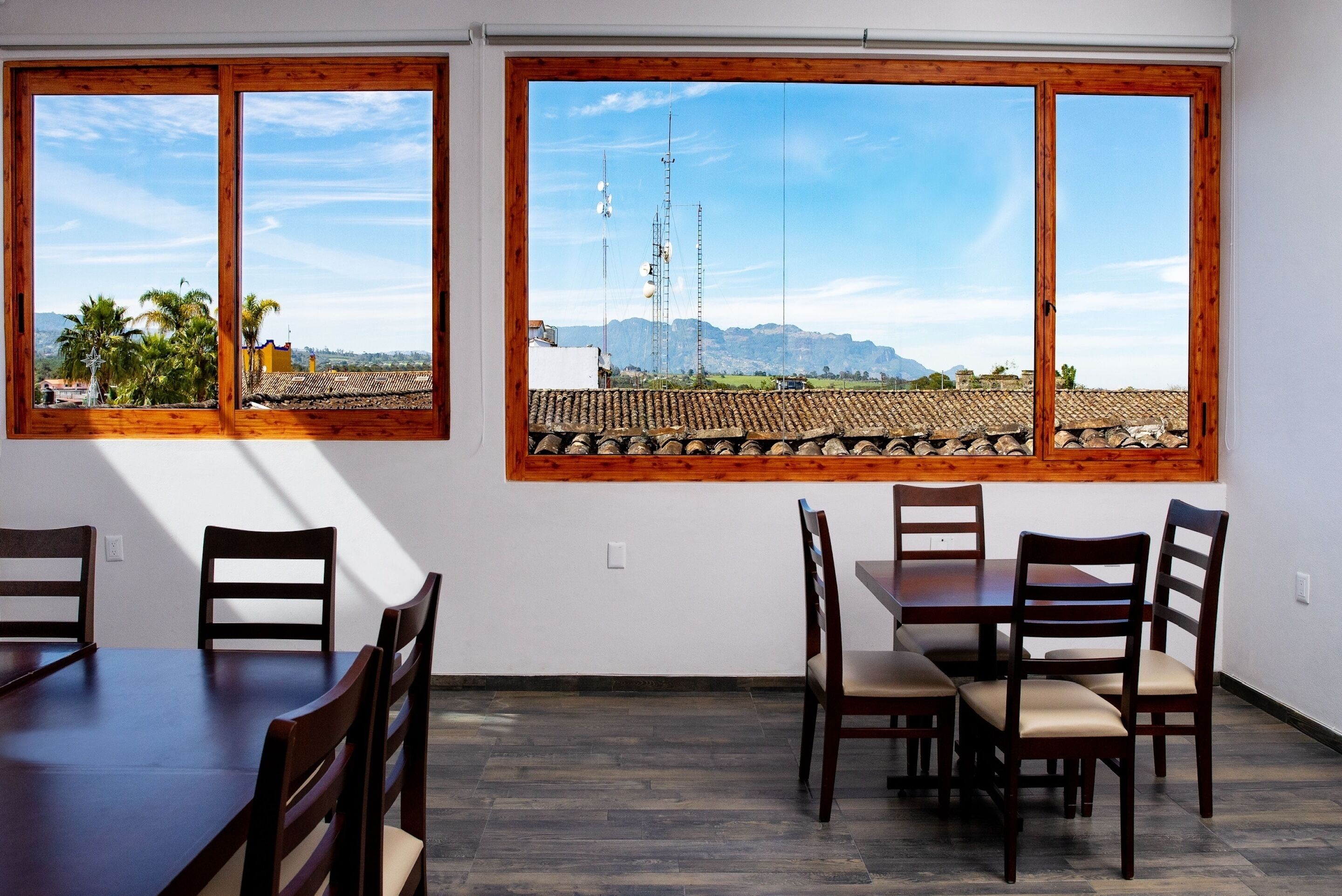 A dining area with a large table and chairs, two windows with wooden frames, and a view of mountains and buildings.