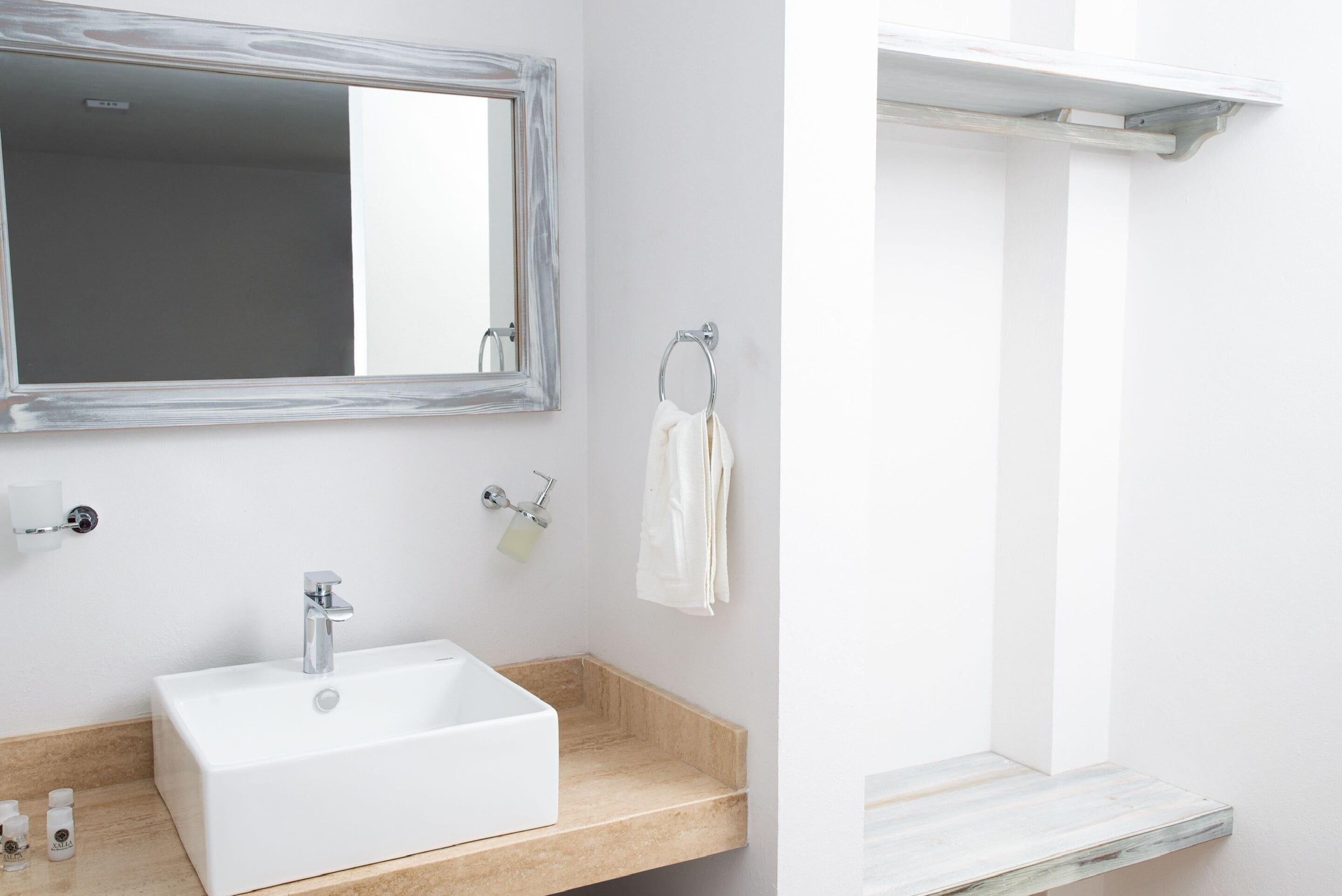 A bathroom with a rectangular sink, a mirror with a wooden frame, a towel rack, and a small shelf.