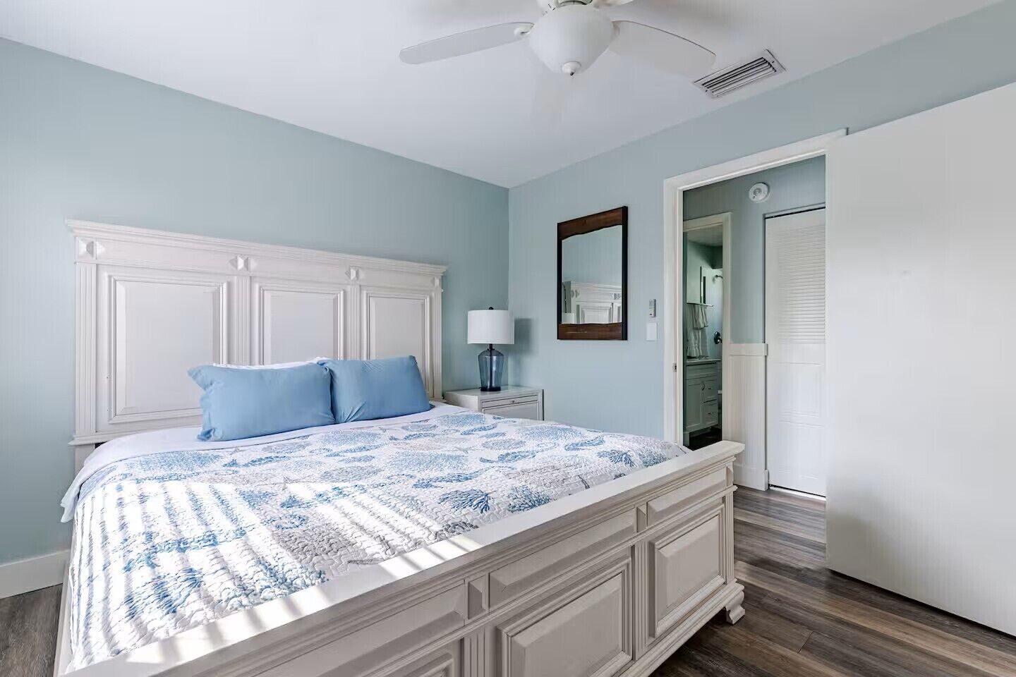 A bedroom with a bed, a nightstand, a ceiling fan, and a door leading to a bathroom.