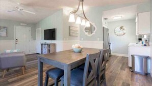 A modern kitchen with a dining area, white cabinets, a ceiling fan, and a wall-mounted TV. - Indian Rocks Gem: Walk to Beach and Dining from Cozy retreat! (Indian Rocks Beach)