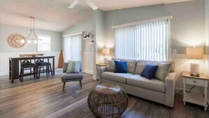 A spacious living room with a sofa, a round wicker coffee table, and a dining area with a table and chairs. - Indian Rocks Gem: Walk to Beach and Dining from Cozy retreat! (Indian Rocks Beach)