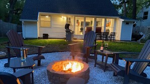Ein Feuerkorb, umgeben von Adirondack-Stühlen, einem Grill und einem Patio-Tisch mit Flasche und Glas.