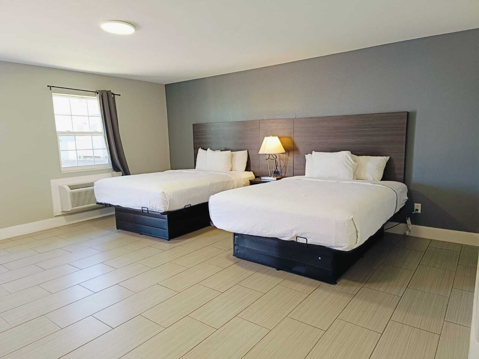 Habitación de hotel con dos camas, una ventana con cortinas, un calentador en la pared y una mesita de noche con lámpara.