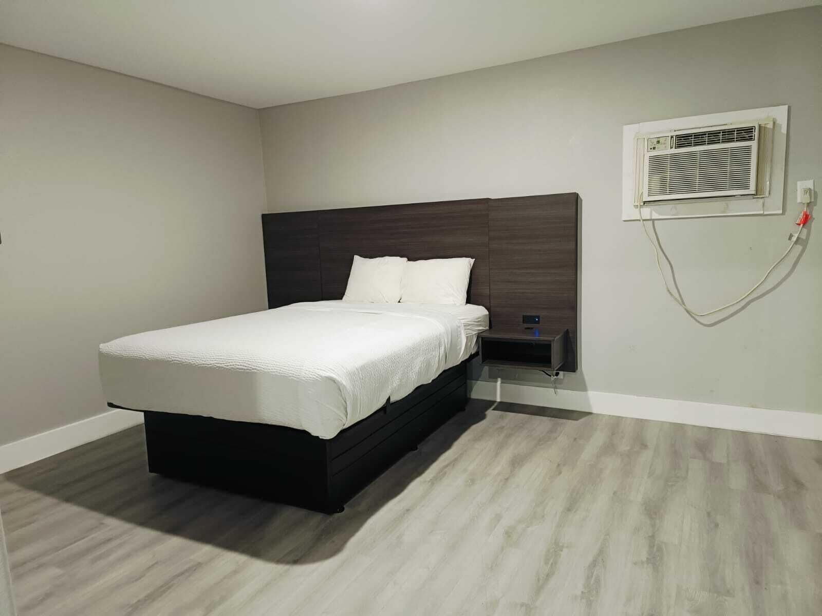 A modern bedroom with a large bed, a nightstand, a wall-mounted air conditioning unit, and a light switch.