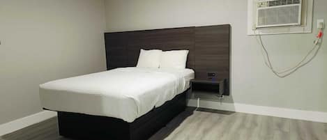 A modern bedroom with a large bed, a nightstand, a wall-mounted air conditioning unit, and a light switch.