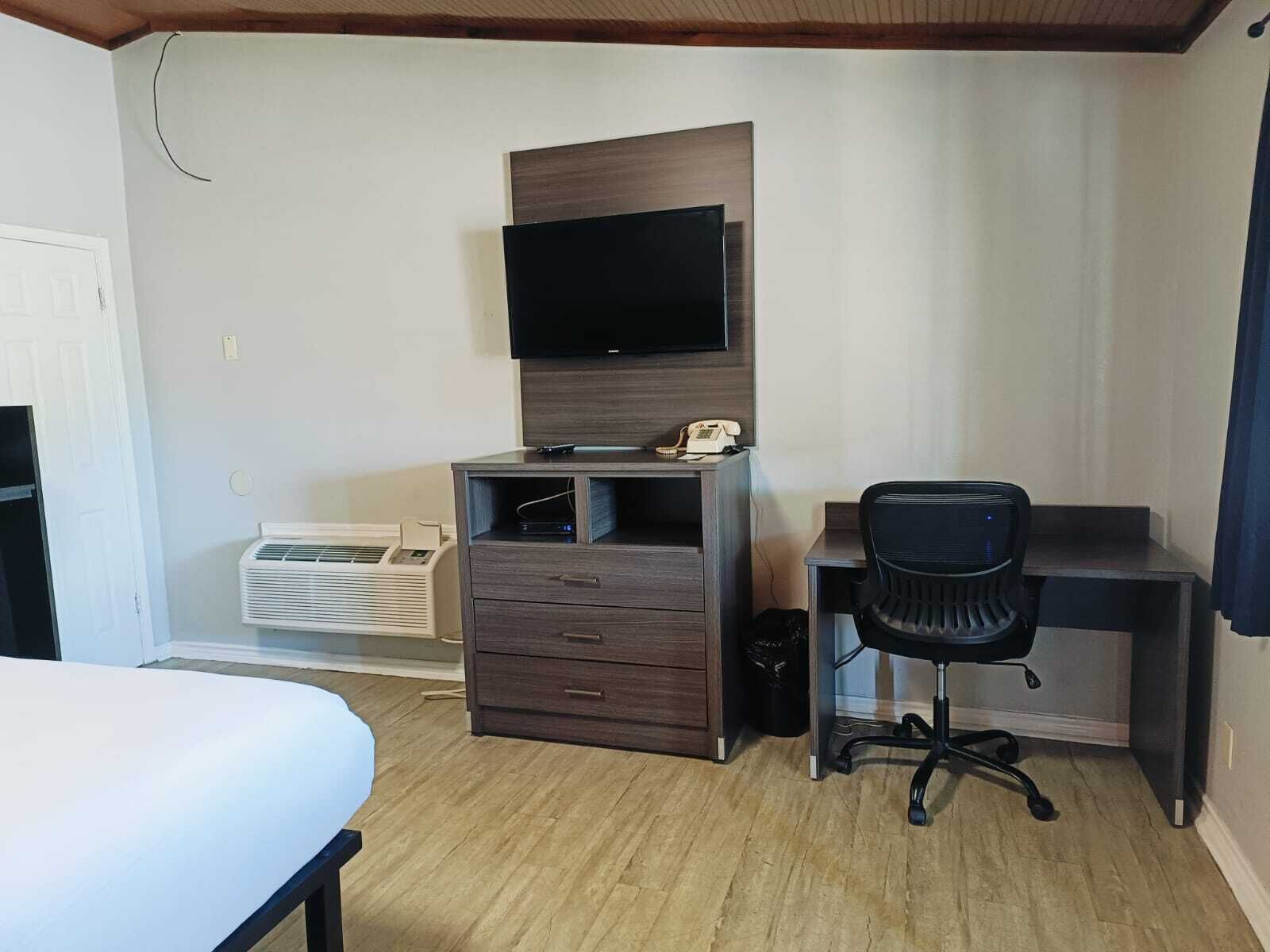 Een compacte hotelkamer met een houten bureau, een zwarte bureaustoel, een flatscreen televisie bevestigd aan een houten paneel, een klein wit nachtkastje en een witte airconditioner.