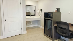 A compact hotel room with a white door, a sink with a mirror, a microwave, a small refrigerator, and a desk with a chair. - Village Inn Motel (Rockport)