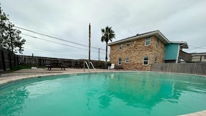 A swimming pool with a stone deck, a wooden fence, and a building with a stone facade and a blue door. - Village Inn Motel (Rockport)