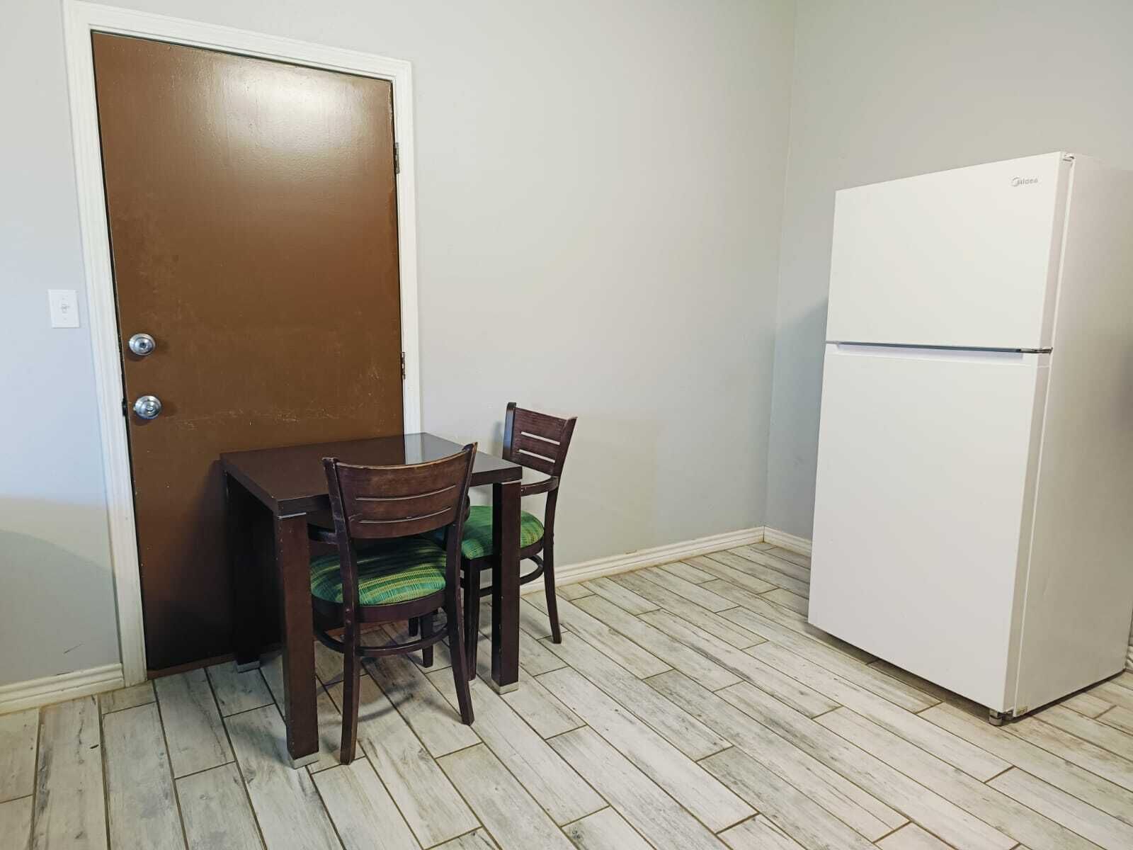 Ein kleiner Raum mit einem Holztisch, zwei Stühlen, einem weißen Kühlschrank und einer braunen Tür.