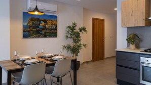 A modern kitchen with a dining area, featuring a wooden table set for two, a potted plant, and a painting of a coastal scene. - Portside Lodge Building (Marsaskala)