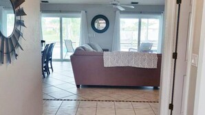 A hallway leading to a living area with a sofa, a ceiling fan, and a view of the outdoors through large windows.