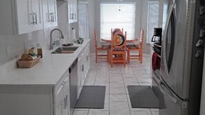A modern kitchen with white cabinets, a stainless steel refrigerator, a dining table with chairs, and a window with blinds.