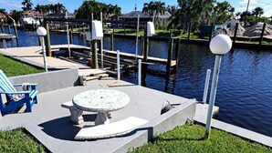 A waterfront patio with a round table and chairs, a lamp post, and a dock extending into the water.