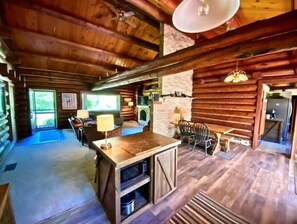 A log cabin interior with a central dining area, a fireplace, and a kitchen area.