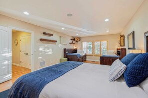 Une chambre avec un grand lit, un bureau et une chaise. Il y a une fenêtre avec des stores, une porte et une horloge au mur.